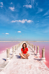 A cute young woman is sitting on a salty beach between wooden sticks on a salty pink lake with a blue sky. A peaceful landscape and relaxation