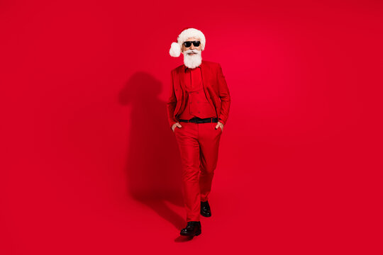 Photo of confident handsome santa claus hands pockets wear sunglass x-mas hat suit shoes on red color background