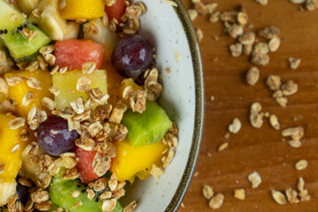 fruit salad with natural cereals served on white dinnerware, cereals scattered around on the table, wooden background in close-up photo taken from above with space for text beside