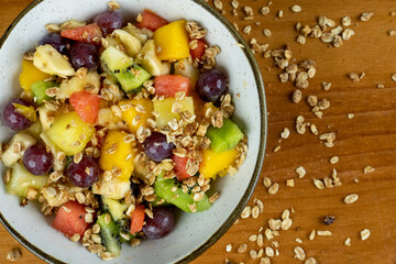 fruit salad with natural cereals served in white dish, cereals scattered around on the table, wooden background in close-up photo taken from above with space for text beside