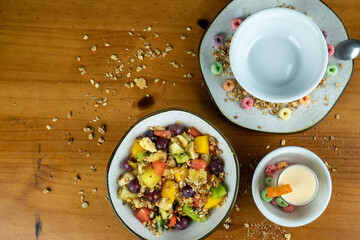 fruit salad with natural cereals served in white dish, decorated with colored cereals, yogurt cup decorated with small orange and cereals spread around on wooden table in photo taken from above.