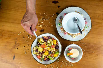 man mixing fruit salad with silver spoon, in white bowl, decorated with colored cereals, and yogurt and cereal spread around on wooden table in photo taken from above.