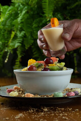 tropical fruit salad served in white bowl on wooden table and green background, decorated with colorful cereal around, man hand holding tilted yogurt cup and decorated with small orange.