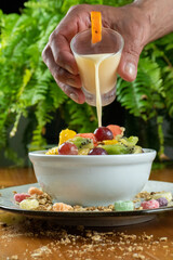 tropical fruit salad on wooden table and green background, decorated with colorful cereal, man pouring yogurt on top of grape, cup decorated with small oranges.