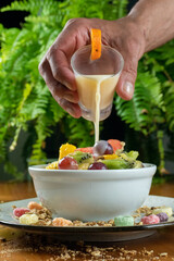 tropical fruit salad on wooden table and green background, decorated with colorful cereal, man pouring yogurt on top of grape, cup decorated with small oranges.
