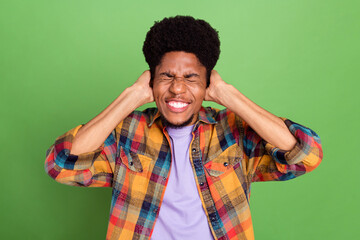 Photo of annoyed unhappy dark skin man wear plaid shirt arms cover ears closed eyes isolated green...