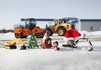 small toy excavators, souvenir snowman, santa claus against the background of large construction equipment