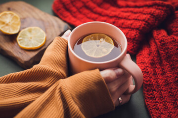 Cup of hot tea with lemon in woman's hands holding it over wooden autumn background with leaves and plaid Top view. Warm drink concept.