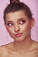 Fashion creative make up. Young woman face expression with clean fresh skin and bright make up with rhinestones freckles on her face posing on a bright pink background