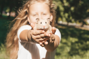 Girl with a hamster in nature. Cheerful happy child girl with pet hamster plays in the backyard of the house in summer. Love, care, tenderness concept.