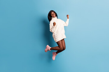 Full length photo of lucky dark skin lady wear white winter outfit jumping high rising fists isolated blue color background