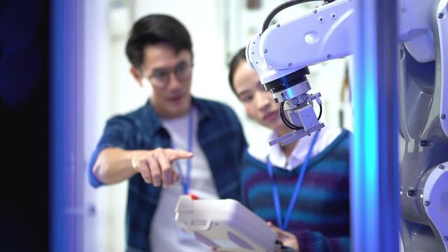 Asian female apprentice engineer in student uniform testing the work of an automation robotic arm machine with a robot controller taught by a young Asian male CEO standing next to her in the factory.
