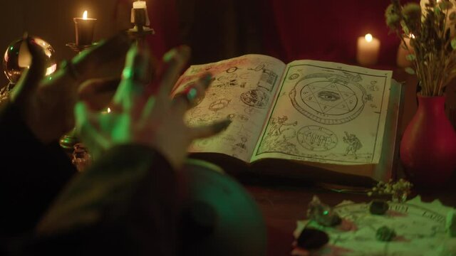 Witch cooking potion elixir close-up. Table with spellbook for making rite. Herbs, burning candles, crystal magic ball for sacrifice. Halloween theme, full moon, witchcraft. 