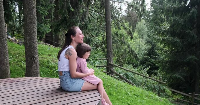 Young woman talking with her daughter while sitting on wooden bench and admiring trees in forest