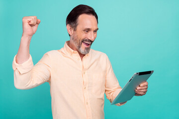 Photo of cheerful smiling handsome mature businessman raise fist in victory hold tablet isolated on turquoise color background