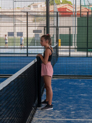 A beautiful young Caucasian teenage girl standing leaning on the net of a paddle tennis court