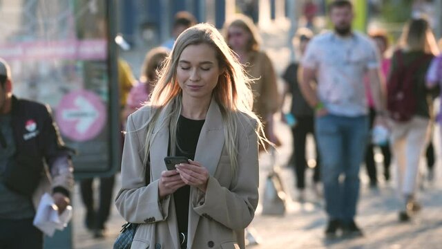 Charming businesswoman, casual looking trendy lady texts on mobile phone. Natural blond woman typing messages on mobile phone touch screen on urban streets background. Portrait of young adult lady.