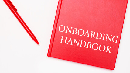 The text ONBOARDING HANDBOOK is written on a red notepad near a red pen on a white table in the...