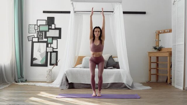 Slender, flexible, caucasian middle-aged woman with black hair in beige tracksuit performs standing asanas Chair Pose, Utkatasana on purple yoga mat in bright room with furniture