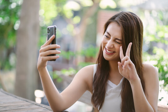 Portrait of cool cheerful Asian girl smile and showing peace sign while taking selfie photo on cellphone in the park. People sincere emotions lifestyle concept.