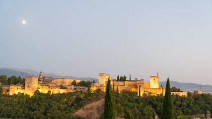 Landscape view of the famous Alhambra illuminated at night, Granada, Andalusia, Spain. White moon on the left. Pale blue sky on the background. With copy-space.