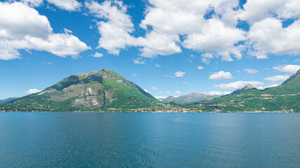 Landscape of the coastline of the famous Lake Como, Italy, with turquoise waters, green hills and Italian alps. In the distance the village of Menaggio is visible. Blue sky and white clouds.