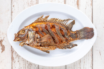 tasty large fried nile tilapia fish in white ceramic plate on white old wood texture background, top view