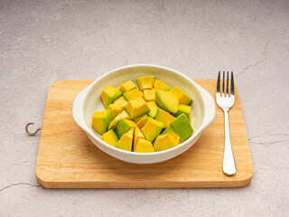 Diced avocados in a ceramic bowl and cutlery on a wooden cutting board on a cement background. Close-up photo. Top view. High vitamin fruit. Healthy fruit concept