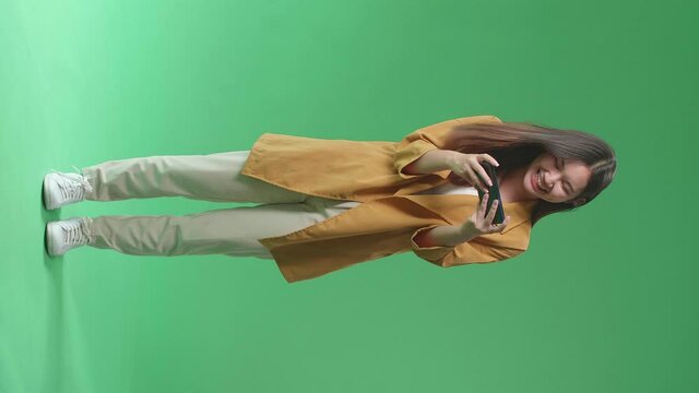 Full Body Of Laughing Asian Woman Looking At The Phone Screen And Holding It In Her Hands While Standing On Green Screen In The Studio
