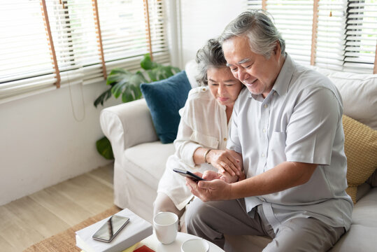 Asian Senior Couple Sitting On Sofa Making Online Video Call With Their Family On Smartphone