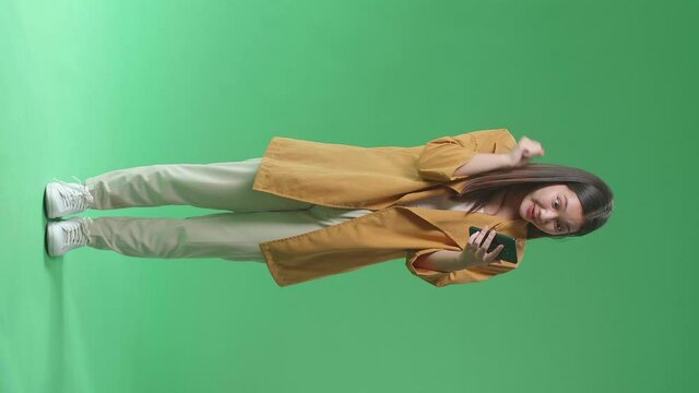Full Body Of Young Asian Woman Flipping Something On The Mobile Phone Becoming Surprised And Opening Her Eyes Wide And Saying Wow While Standing In The Green Screen Studio
