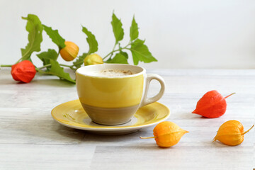 The concept of a good autumn morning. Yellow cup of coffee with branches of physalis on a gray table, side view, space for text
