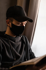 Fototapeta na wymiar young man with protective medical mask and black cap in hoodie reading magazine indoors