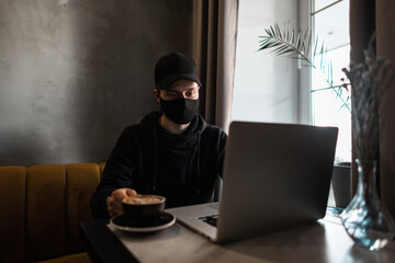 Young guy with a medical protective mask and a black cap in a hoodie drinks coffee and looks at a laptop in a cafe