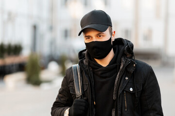 Fashionable handsome guy with a protective mask and a black cap in a stylish hoodie and jacket with a backpack walks on the street. COVID-19 and pandemic concept