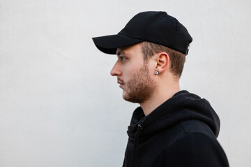 Handsome young man with stubble in a fashionable black hoodie with a stylish black cap near a white...