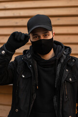 Portrait of a handsome young guy with a protective medical mask in a black jacket with a hoodie straightens his cap on a wooden background