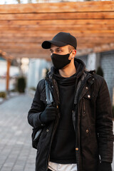 Stylish handsome man with a protective mask in fashionable clothes with a jacket, hoodie, mockup cap and backpack walks in the city. Men's urban clothing style