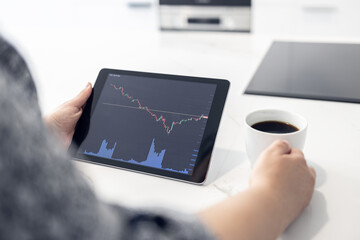 Woman looking at a digital tablet with stock market chart and holding a cup of coffee in a modern kitchen. Trading at home concept