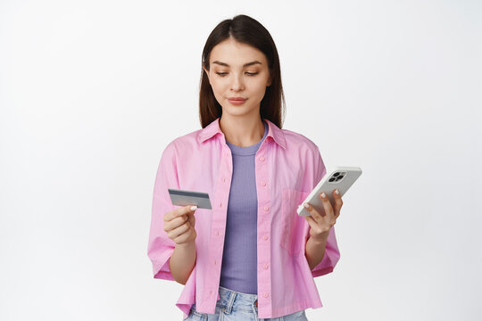 Image of smiling woman making purchase on mobile phone, looking at credit card while order something on smartphone app, standing over white background