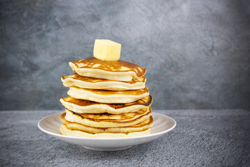 Stack of pancakes on white plate with piece of butter on top, on gray surface