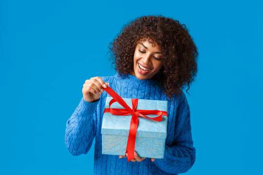 Curious And Happy, Smiling African-american Woman, B-day Girl In Winter Sweater, Pulling Present Knot To Unwrap Gift And See What Inside, Celebrating Christmas, New Year Holidays, Blue Background