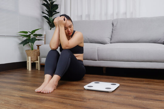 Young Asian woman sitting on the floor looking at the weight scale worried and depressed in dieting and eating disorder concept