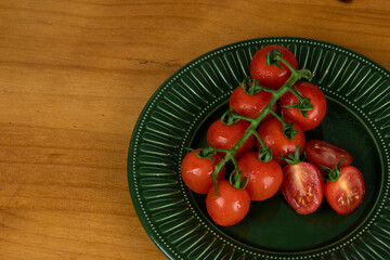 beautiful small tomatoes, whole and sliced, with drops of water, green leaves served in dark green plate on wooden table and light background, photo from above, space in the background for text