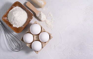 The flour is in a wooden bowl, the eggs are lying on a bright table. Rolling pin and whisk in the background. Space for text