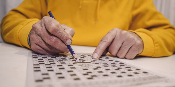 Male wrinkled hand holding blue pencil and writing letter in crossword