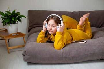 cute little girl in yellow clothes lies on the bed and listens to music, watches videos and communicates on social networks through a smartphone with large white headphones. modern technologies
