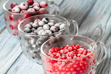 Ripe frozen sweet cherries, red currant and black currant with hoarfrost in the transparent glass cups on blue wooden background. Natural organic healthy food. Closeup, selective focus