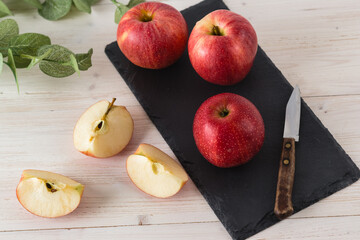 In a rectangular black slate dish on white wooden table are three juicy beautiful ripe red apples of the early winter variety Gala. Next to the slices of the cut fruit and a knife. Top view