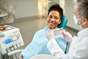 Happy black woman and her dentist choosing the shade of dental veneers during appointment at dental...